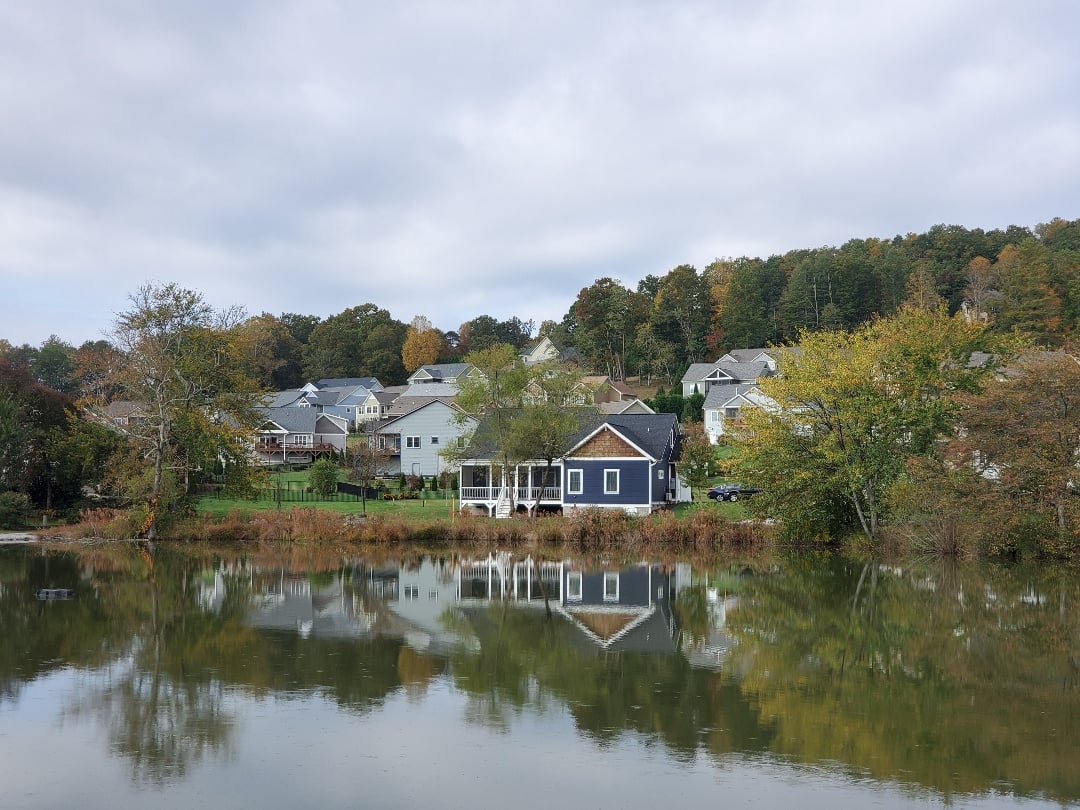 A home in a neighborhood rests next to a glassy lake with cloudy skies overhead.