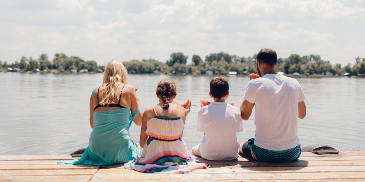 A family of four sits on a dock eating watermelon while looking over the lake.